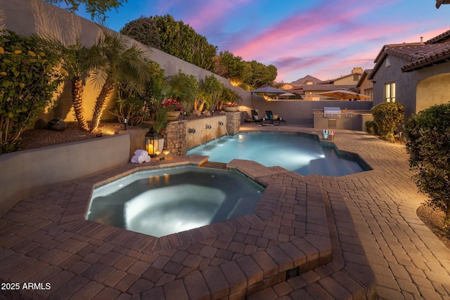 pool at dusk featuring an in ground hot tub, pool water feature, exterior kitchen, and a patio area