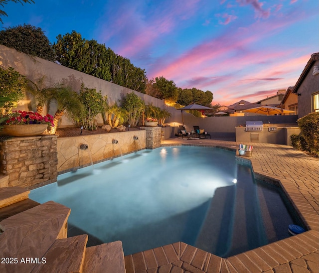 pool at dusk featuring pool water feature, grilling area, exterior kitchen, and a patio