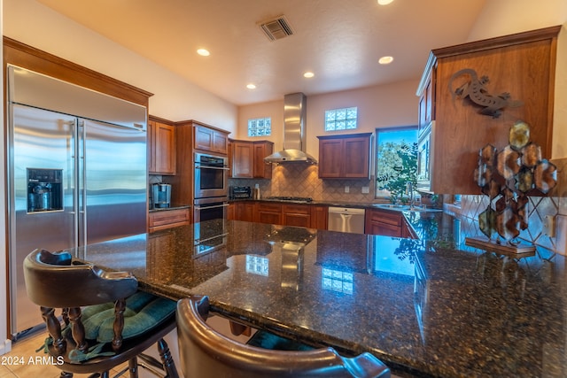 kitchen with sink, wall chimney exhaust hood, stainless steel appliances, a kitchen breakfast bar, and dark stone countertops