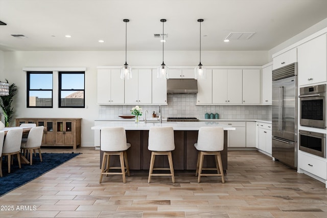 kitchen featuring appliances with stainless steel finishes, backsplash, white cabinets, a center island with sink, and decorative light fixtures