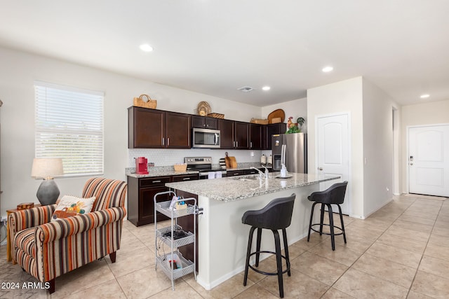 kitchen featuring a center island with sink, appliances with stainless steel finishes, dark brown cabinets, light stone counters, and a breakfast bar area