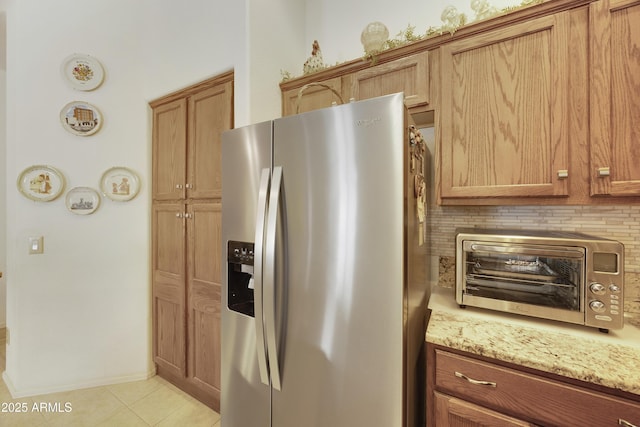 kitchen featuring stainless steel fridge, light tile patterned flooring, light stone counters, brown cabinets, and backsplash