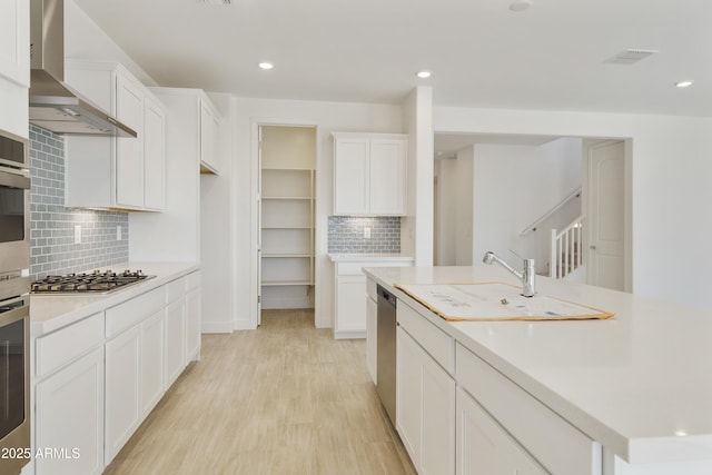 kitchen with white cabinets, light hardwood / wood-style flooring, wall chimney exhaust hood, tasteful backsplash, and stainless steel appliances