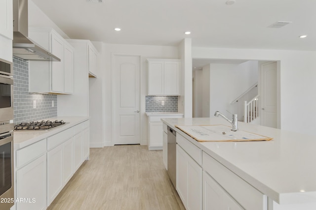 kitchen featuring appliances with stainless steel finishes, backsplash, wall chimney range hood, light hardwood / wood-style flooring, and white cabinets