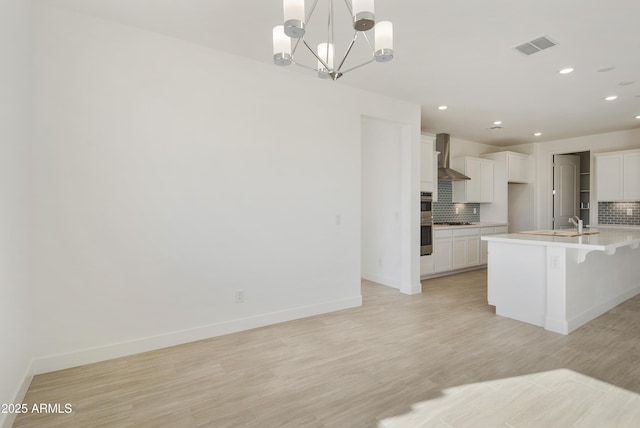 kitchen featuring wall chimney exhaust hood, decorative backsplash, an island with sink, and a notable chandelier