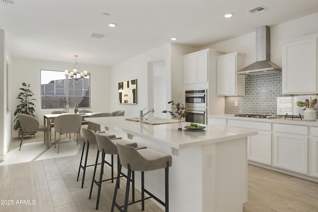 kitchen with stainless steel appliances, white cabinets, wall chimney exhaust hood, and an island with sink