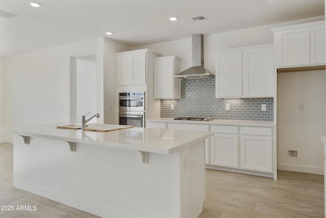 kitchen featuring wall chimney exhaust hood, stainless steel appliances, sink, a center island with sink, and white cabinetry