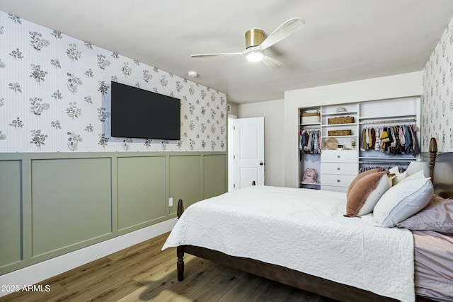 bedroom with wood finished floors, wallpapered walls, a closet, a wainscoted wall, and a ceiling fan