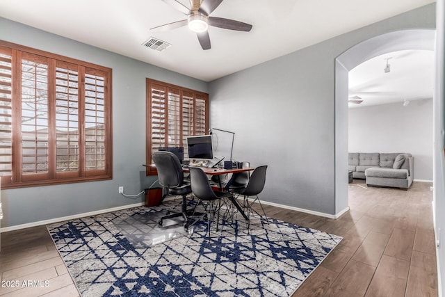 home office featuring arched walkways, wood finished floors, a ceiling fan, visible vents, and baseboards