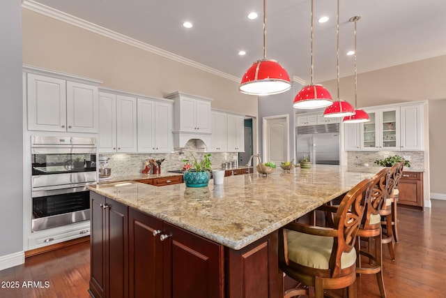 kitchen with glass insert cabinets, dark wood finished floors, crown molding, stainless steel appliances, and a large island with sink