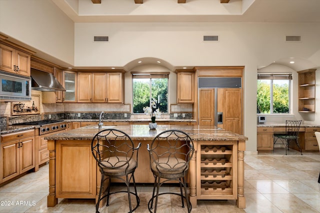 kitchen with built in appliances, stone counters, a center island with sink, and wall chimney range hood