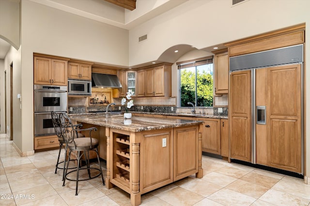 kitchen featuring backsplash, built in appliances, wall chimney exhaust hood, dark stone countertops, and an island with sink