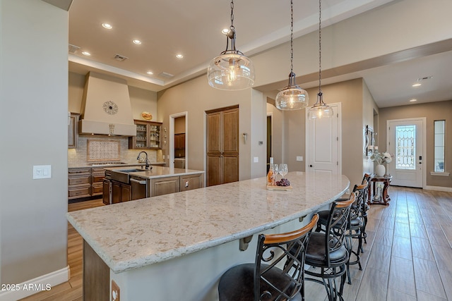 kitchen with premium range hood, sink, tasteful backsplash, a center island with sink, and pendant lighting