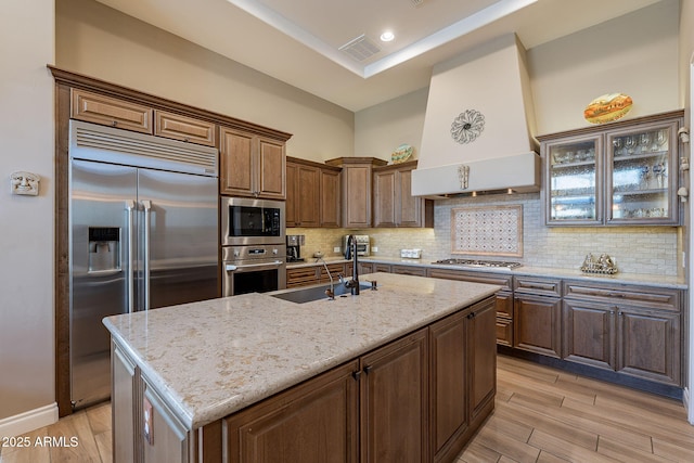 kitchen featuring sink, decorative backsplash, a kitchen island with sink, built in appliances, and custom range hood