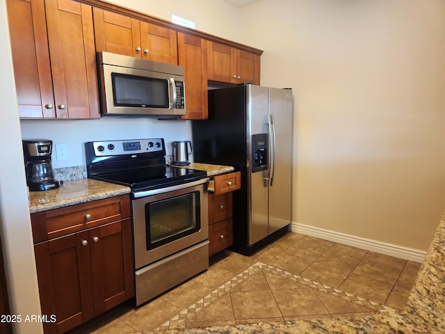 kitchen with brown cabinets, appliances with stainless steel finishes, light tile patterned floors, baseboards, and light stone countertops