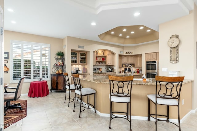 kitchen with a kitchen bar, a kitchen island with sink, a tray ceiling, light stone counters, and stainless steel double oven