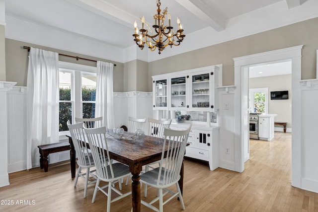 dining space featuring wainscoting, beamed ceiling, light wood-type flooring, a decorative wall, and a notable chandelier