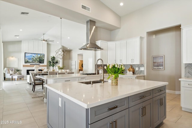 kitchen featuring a center island with sink, tasteful backsplash, gray cabinetry, light tile patterned flooring, and wall chimney range hood