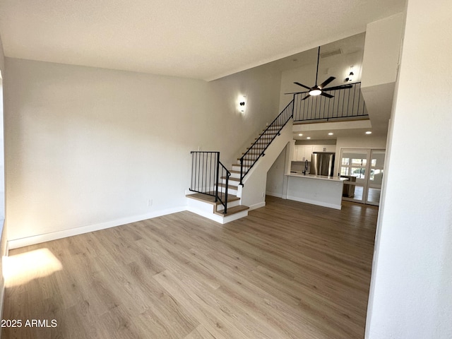 unfurnished living room with stairs, a towering ceiling, light wood-style floors, baseboards, and a ceiling fan