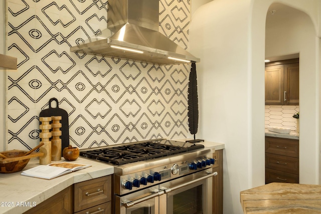 kitchen with arched walkways, backsplash, range with two ovens, light stone counters, and under cabinet range hood