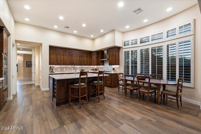 kitchen with tasteful backsplash, wall chimney exhaust hood, dark brown cabinetry, a kitchen island with sink, and dark wood-type flooring