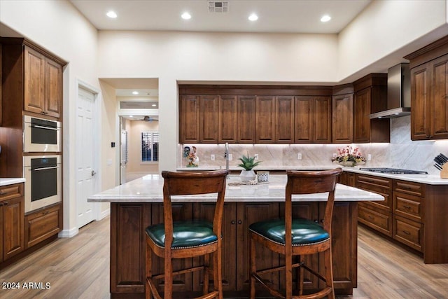 kitchen with a breakfast bar, wall chimney range hood, light wood-type flooring, appliances with stainless steel finishes, and a kitchen island