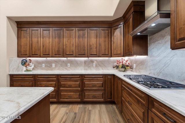 kitchen featuring backsplash, wall chimney range hood, light stone countertops, light hardwood / wood-style floors, and stainless steel gas cooktop