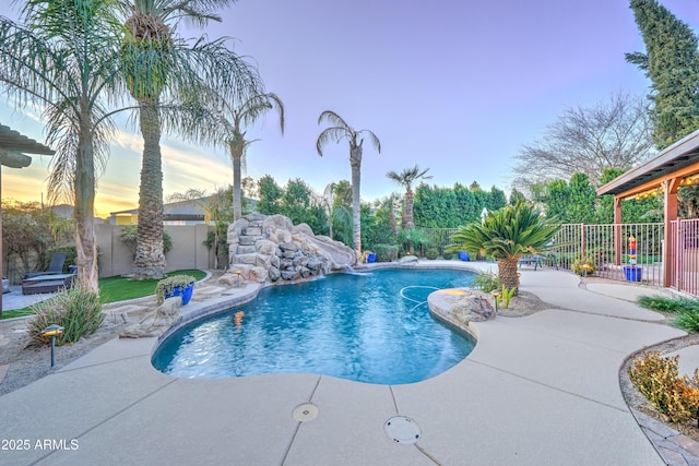 pool at dusk with pool water feature, a water slide, and a patio area