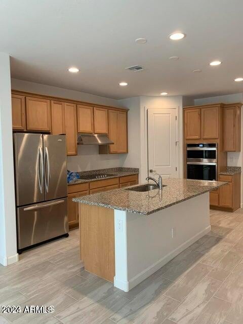 kitchen with light wood-type flooring, light stone counters, sink, an island with sink, and appliances with stainless steel finishes