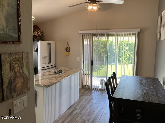 kitchen with white fridge with ice dispenser, ceiling fan, vaulted ceiling, light stone counters, and light wood-style flooring