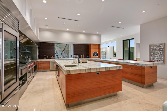 kitchen with range hood, tasteful backsplash, stainless steel built in refrigerator, sink, and a large island with sink