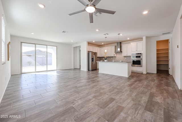 kitchen featuring appliances with stainless steel finishes, white cabinetry, hanging light fixtures, a center island with sink, and wall chimney exhaust hood