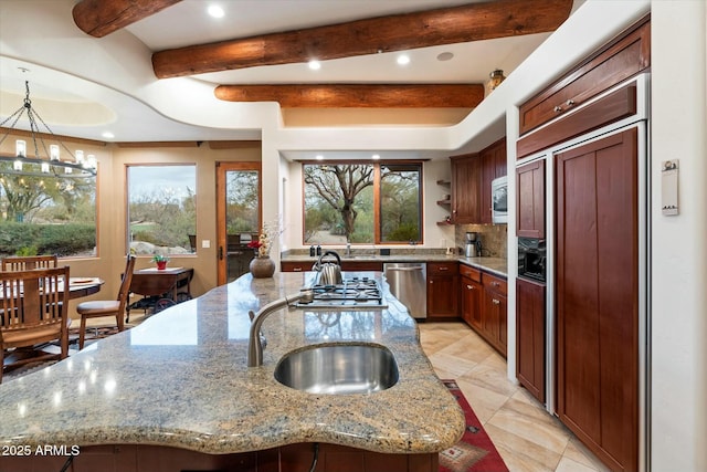 kitchen with stainless steel appliances, light stone countertops, hanging light fixtures, and a center island with sink