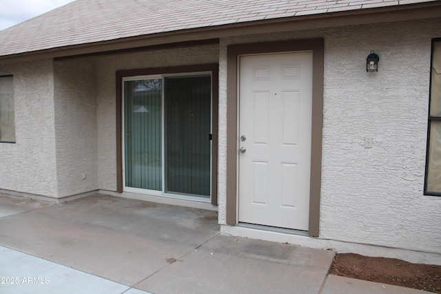 entrance to property with stucco siding, roof with shingles, and a patio area