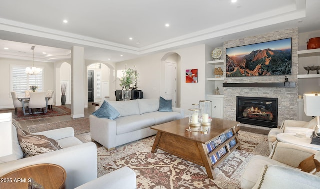 living room with built in shelves, a raised ceiling, a notable chandelier, and a stone fireplace