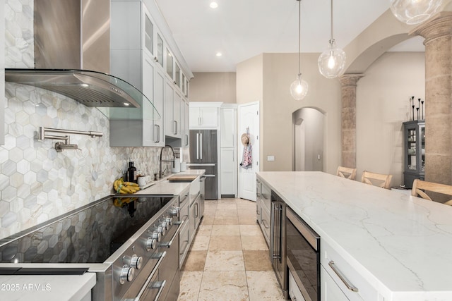 kitchen featuring decorative columns, wall chimney exhaust hood, a sink, backsplash, and stainless steel appliances