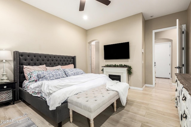 bedroom featuring ceiling fan, light wood finished floors, a fireplace, arched walkways, and baseboards
