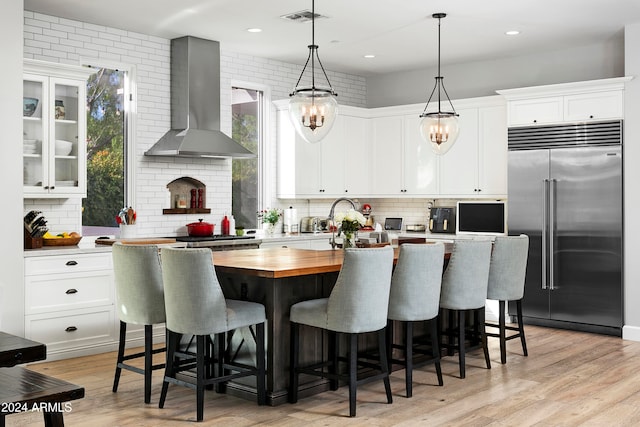 kitchen featuring wall chimney exhaust hood, hanging light fixtures, stainless steel appliances, a center island with sink, and white cabinets