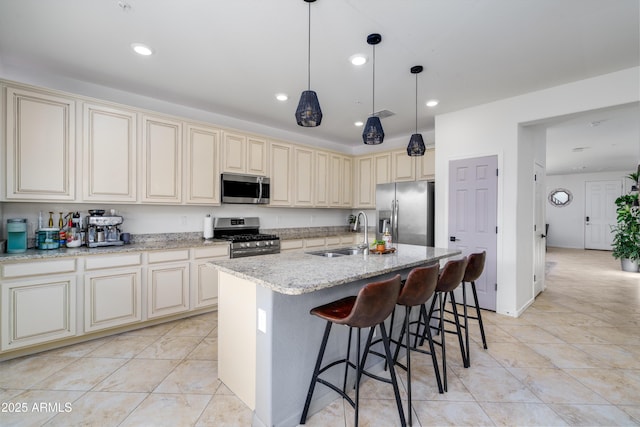 kitchen with sink, appliances with stainless steel finishes, light stone countertops, a center island with sink, and cream cabinetry