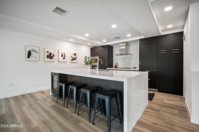 kitchen featuring light wood-type flooring, a large island with sink, sink, wall chimney exhaust hood, and a kitchen breakfast bar