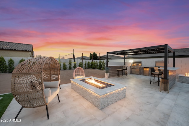 patio terrace at dusk with exterior kitchen, a fire pit, fence, and a grill