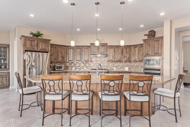 kitchen featuring light stone counters, hanging light fixtures, appliances with stainless steel finishes, a kitchen breakfast bar, and a kitchen island with sink