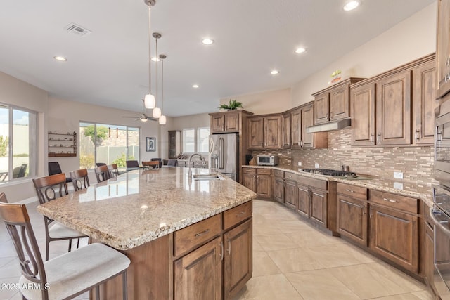 kitchen featuring a kitchen island with sink, hanging light fixtures, stainless steel appliances, and light stone countertops