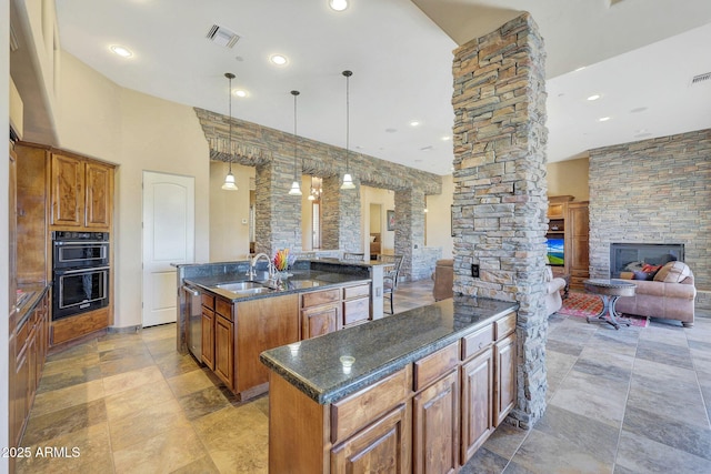 kitchen with decorative columns, black double oven, a fireplace, hanging light fixtures, and an island with sink