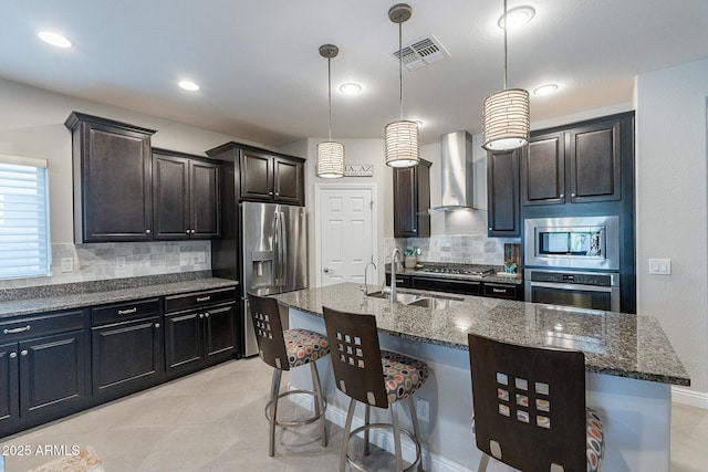 kitchen with pendant lighting, appliances with stainless steel finishes, an island with sink, dark stone counters, and wall chimney exhaust hood