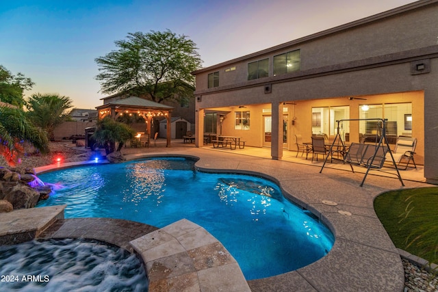 pool at dusk featuring an in ground hot tub, pool water feature, ceiling fan, a gazebo, and a patio area