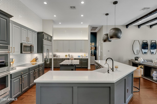 kitchen with beam ceiling, gray cabinetry, stainless steel appliances, decorative light fixtures, and a kitchen island with sink