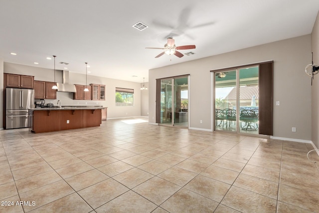 kitchen featuring wall chimney range hood, hanging light fixtures, ceiling fan, stainless steel fridge, and an island with sink