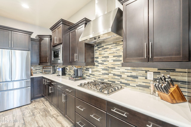 kitchen featuring light countertops, dark brown cabinets, appliances with stainless steel finishes, wall chimney exhaust hood, and backsplash