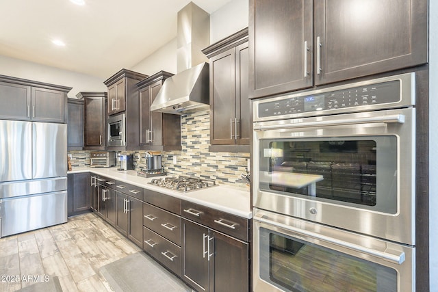 kitchen with dark brown cabinetry, light countertops, appliances with stainless steel finishes, wall chimney exhaust hood, and tasteful backsplash